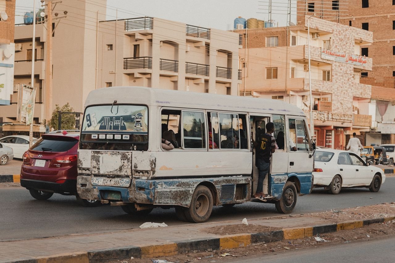 Minibus navigating busy city street with passengers and urban backdrop.