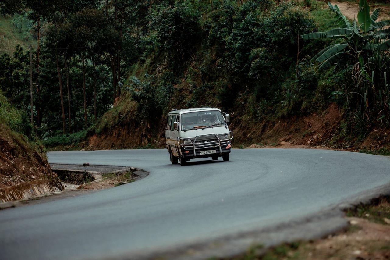 A white minibus drives through a lush, winding rural road surrounded by greenery.