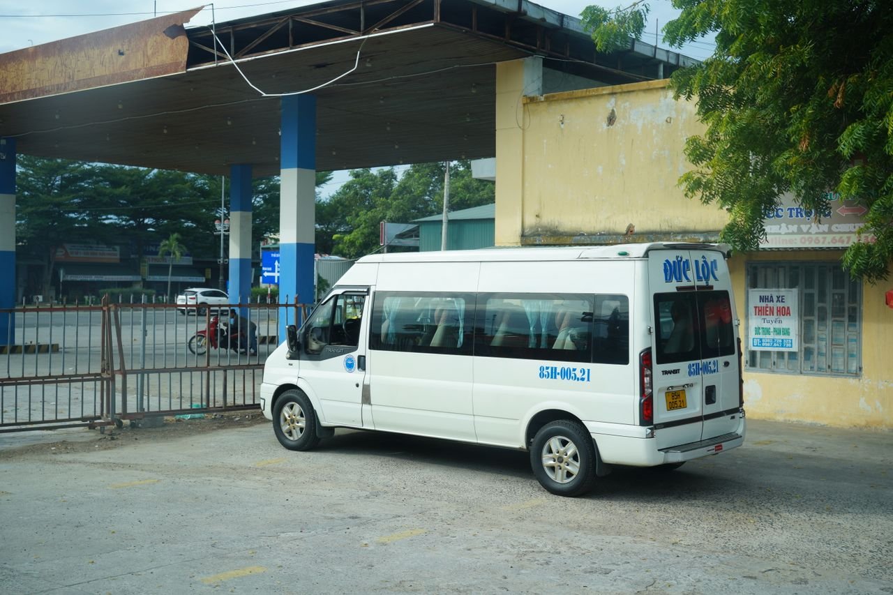 A white minibus is parked at a bus station under a rustic metal canopy.