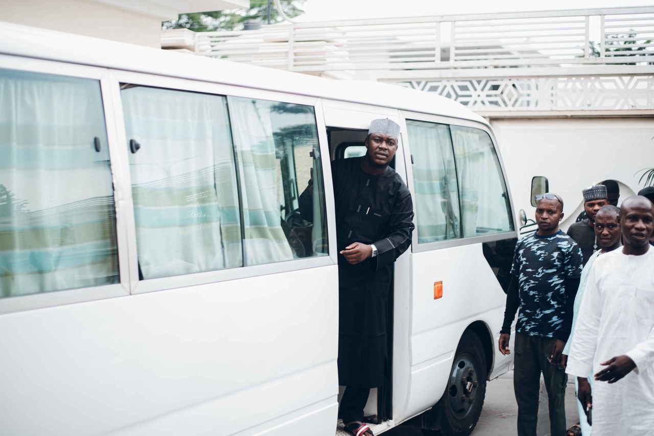 A group of men enter a white minibus parked outdoors, dressed in various attires.