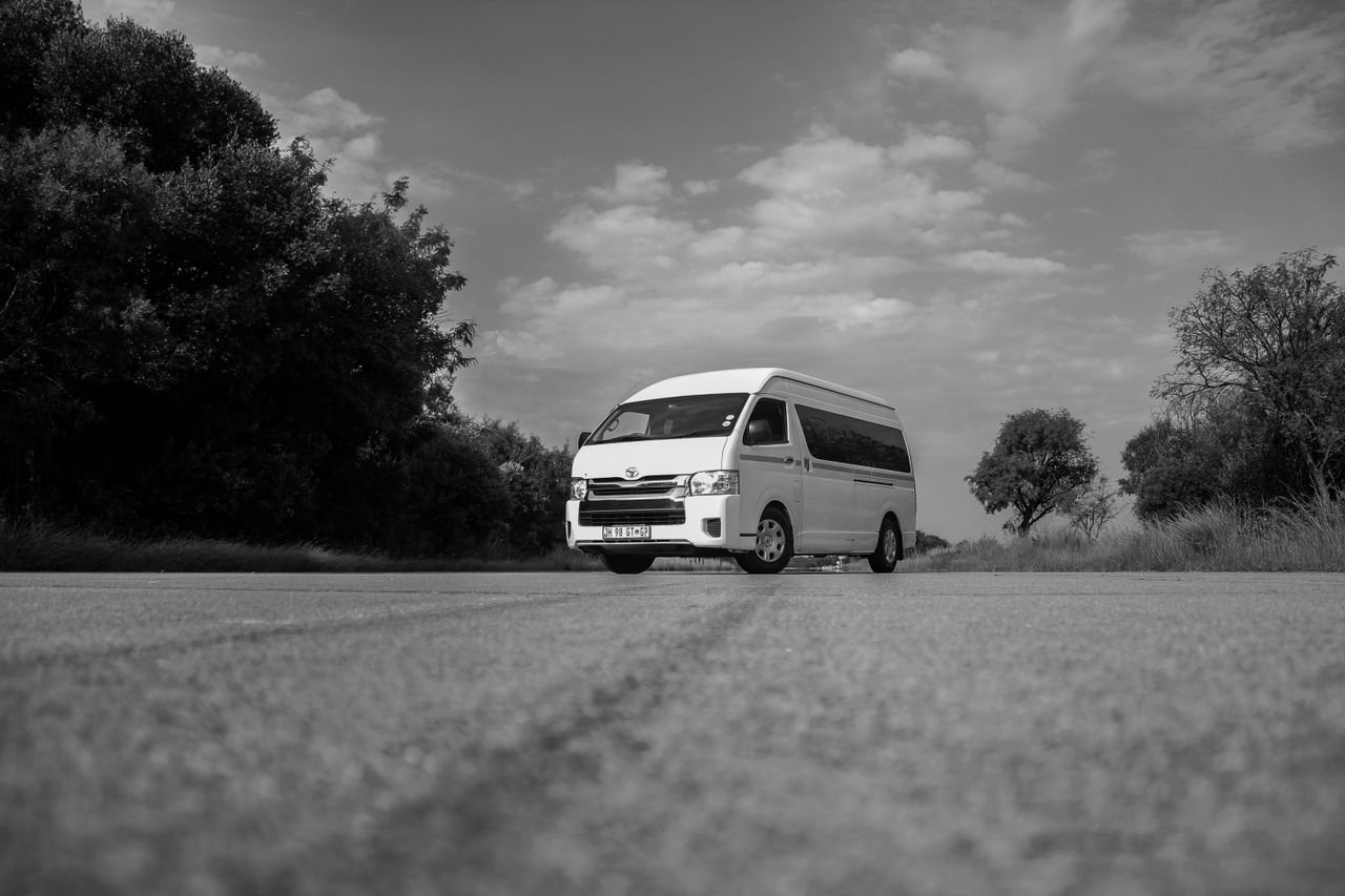 Black and white photo of a white van driving on an empty countryside road under a cloudy sky.